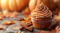 Pumpkin spice cupcake with cinnamon frosting swirl on rustic wooden background with autumn leaves and cinnamon sticks, warm fall colors and bokeh lighting.
