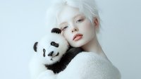 Young woman with albinism features and white hair holding plush panda toy against white background, creating ethereal artistic portrait with soft lighting and minimalist style.