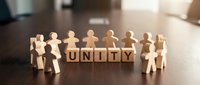 Wooden figures holding hands in circle around unity text blocks on table, representing teamwork, cooperation, and community togetherness.