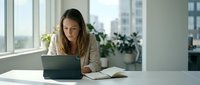 Young professional woman working on laptop at modern office desk with notebook and plants by window during daytime business hours.