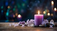 Purple candle burning surrounded by lilac flowers and crystals on wooden surface with colorful bokeh lights creating peaceful spa atmosphere for relaxation.