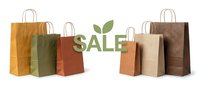 Eco-friendly kraft paper shopping bags in earthy tones arranged around a green sale sign with leaves, isolated on white background.