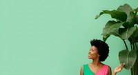 Woman with dark curly hair and medium skin tone wearing colorful green and pink sleeveless top, posing next to large potted plant against mint green background for lifestyle and wellness content.