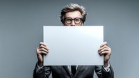 Senior woman with glasses peeking over blank white poster against gray background, perfect for adding custom message or advertisement.