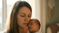 Young Caucasian mother holding sleeping newborn baby in tender embrace by window with soft natural light creating intimate bonding moment for family content.