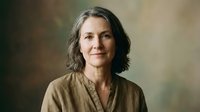 Middle-aged woman with long dark hair and fair complexion wearing a neutral beige linen shirt, professional portrait against soft blurred background for corporate, business, or editorial use.