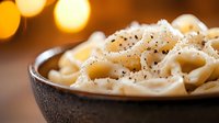 Creamy macaroni pasta with black pepper in dark ceramic bowl against warm bokeh lights background, comfort food close-up with selective focus.