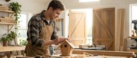 Caucasian male carpenter in plaid shirt and apron working with wood in bright workshop, sanding or finishing wooden surface at workbench with tools and materials.