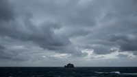 Dramatic stormy sky with dark clouds over turbulent ocean waters and distant rocky island formation on horizon.