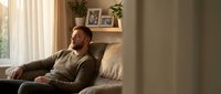 Middle-aged man with dark beard and South Asian appearance sitting on beige sofa in modern living room with natural light from window, relaxed home lifestyle setting.