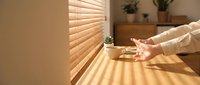 Woman adjusting wooden blinds in bright sunlit room with natural light streaming through window creating shadow patterns on table surface.