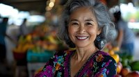 Smiling mature Asian woman with gray hair wearing colorful floral blouse at farmers market with fresh produce background for lifestyle and wellness content.