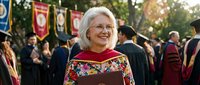 Senior Caucasian woman in colorful graduation regalia smiling at outdoor university commencement ceremony with academic banners and crowd in background.