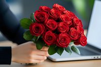 Vibrant red roses bouquet held by woman's hands near laptop, fresh romantic flowers with green leaves against blurred office background.