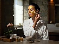 Young woman applying skincare serum in luxury spa bathroom wearing white robe with natural lighting and organic beauty products on wooden table.