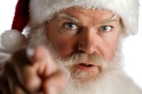 Close up portrait of mature bearded man in Santa hat with stern expression pointing finger at viewer, dramatic lighting highlights facial features and white beard.