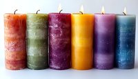 Colorful rustic pillar candles in rainbow order - orange, green, burgundy, yellow, purple and blue - lined up with soft glowing flames on white background.