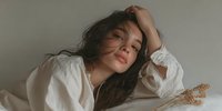 Young Hispanic woman with long dark hair resting hand on forehead in white shirt, expressing fatigue or headache in soft natural lighting.