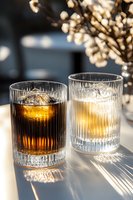 Two elegant cocktails in crystal ribbed glasses with spherical ice, backlit by warm sunlight casting dramatic shadows. Amber and clear spirits create luxurious mood.
