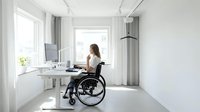 Young woman in wheelchair working at modern white desk in bright minimalist office space with large windows and contemporary design for accessibility.