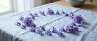 Purple paper flowers arranged in heart shape on white linen fabric creating romantic flat lay composition for wedding or Valentine's Day crafts.