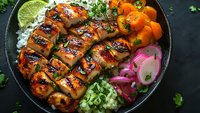 Grilled teriyaki chicken breast sliced and served with steamed rice, pickled red onions, fresh herbs and glazed carrots in black bowl on dark background.
