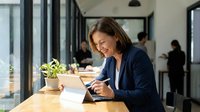 Professional middle-aged Caucasian businesswoman smiling while using tablet in modern office workspace with natural lighting and colleagues in background.