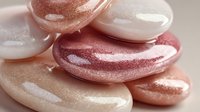 Shimmering smooth pebbles in soft pink, peach, and white tones stacked together with glossy, metallic finish on neutral background.