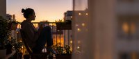 Young woman relaxing on urban balcony at sunset with string lights and plants creating cozy evening atmosphere for home lifestyle content.