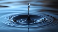 Water drop collision creating perfect concentric ripples on calm blue surface with secondary droplet suspended above in high-speed macro photography.