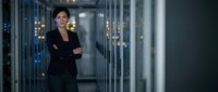 Professional Asian businesswoman standing confidently in modern data center server room with arms crossed wearing black suit for corporate technology concepts.