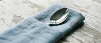 Vintage silver spoon resting on folded blue linen napkin against weathered white wooden surface for dining and tableware design projects.