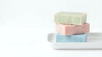 Stacked natural soap bars in pastel colors including green, pink, and blue on white ceramic dish against neutral background for spa and wellness design.