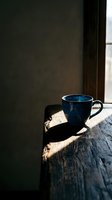 Blue ceramic mug on rustic wooden table with natural sunlight streaming through window, creating dramatic shadows. Minimalist still life composition for beverage, lifestyle, and interior design projects.