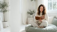 Young Caucasian woman with curly hair reading a book on a cozy floor cushion in a bright minimalist living room with plants.
