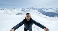 Man in black winter jacket with arms outstretched standing on snowy mountain peak with alpine landscape backdrop, expressing joy and freedom during winter adventure travel.