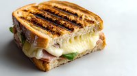 Grilled sandwich with melted cheese and ham on toasted white bread, served on white background, close up view showing grill marks and oozing cheese.