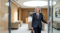 Professional mature blonde businesswoman in dark suit opening glass office door in modern corporate lobby with wooden panels and contemporary furniture.