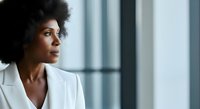 Thoughtful African American woman in white blazer gazing out window in modern office setting, suitable for corporate, business, and professional lifestyle content.
