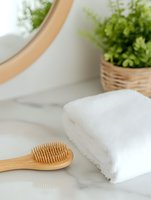 Soft white towel and bamboo hairbrush on marble surface with green plant and mirror in bathroom setting, creating serene spa atmosphere.