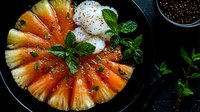 Fresh pineapple slices arranged in circle with mint leaves and spices on dark plate, top view. Juicy tropical fruit with chili flakes for sweet and spicy dish.