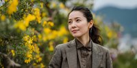 Professional Asian businesswoman in brown suit smiling outdoors with yellow flowers in spring garden background for corporate lifestyle content.