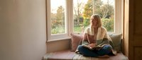 Young blonde woman in cozy sweater reading book by window seat with autumn park view. Peaceful indoor lifestyle moment for relaxation and learning concepts.