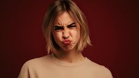 Young Asian woman with blonde bob haircut making funny face with puffed cheeks and furrowed brows against dark red background, expressing playful emotion.