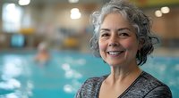 Mature woman with natural gray curly hair smiling at indoor swimming pool, wearing gray athletic wear. Active senior enjoying water fitness and healthy lifestyle.