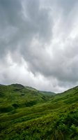 Dramatic stormy sky looms over lush green rolling hills and mountains covered in vegetation, creating moody landscape scenery for nature and weather projects.