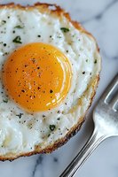 Close-up of crispy fried egg with bright yellow yolk, seasoned with black pepper and herbs on marble surface, fork visible in corner. Breakfast food photography.