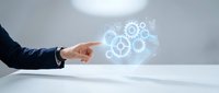 Businessman in dark suit pointing at glowing gear mechanism hologram floating above white desk, representing digital technology, automation, and business process optimization.