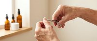 Senior man applying skincare serum from dropper bottle in modern bathroom with amber glass containers on shelf for daily grooming routine.