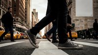 Urban street scene with person walking in sneakers on city crosswalk with yellow taxis and tall buildings in background during daytime rush hour.